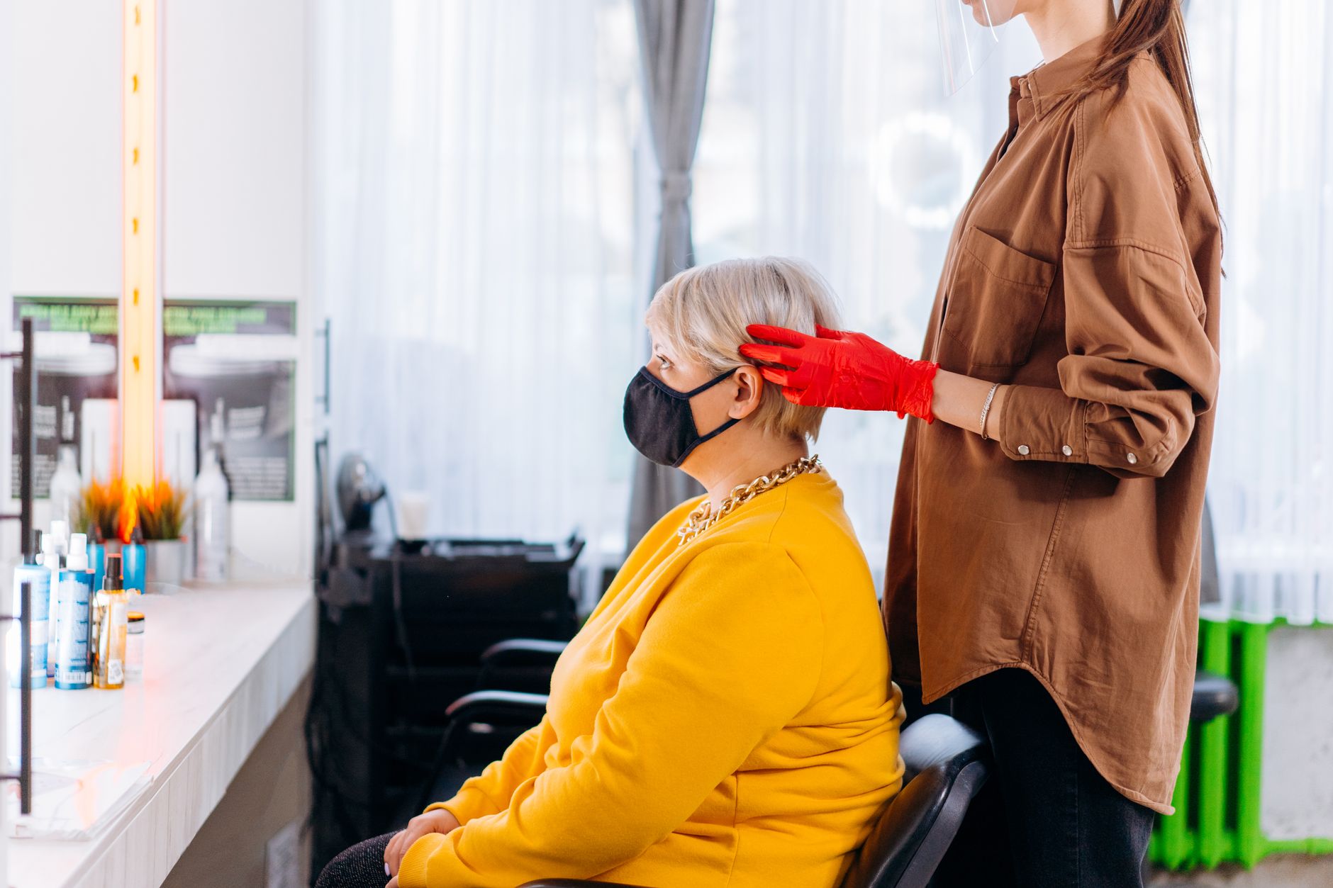 Side view of female client and hairstylist in masks and casual clothes with gloves near table with professional equipment and mirror in bright salon